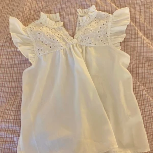 Vit blus med spets och volang - Säljer en söt vit blus med broderad spets upptill och volangdetaljer vid axlar och hals. Blusen är ärmlös och med fina broderier och rynkade detaljer. Perfekt för vår och sommar! 😻