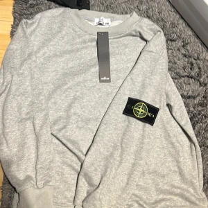 Grå sweatshirt från Stone Island - Säljer en stilren grå sweatshirt från Stone Island med klassisk rund hals och ribbade muddar. Tröjan har den ikoniska Stone Island-loggan på vänster ärm. Perfekt för en avslappnad och trendig look.