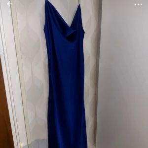 Blå långklänning med smala axelband - Säljer en elegant blå långklänning med smala glittriga axelband och draperad urringning. Klänningen har en slank siluett och är tillverkad i ett glansigt material som ger en lyxig känsla. Perfekt för speciella tillfällen. Aldrig använd, helt ny!