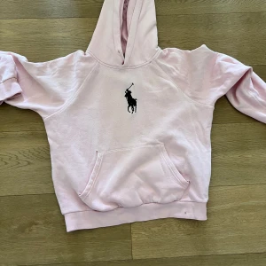 (OBS!)Ljusrosa hoodie från Polo Ralph Lauren - Säljer en ljusrosa hoodie från Polo Ralph Lauren med klassisk logga broderad på bröstet. Tröjan har huva, magficka och ribbade muddar. Perfekt för en avslappnad och stilren look. Jag skrev obs för att det fann två små hål vid magen men det är sytt ihop men där med det billiga priset.