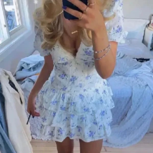 Vit blommig kortklänning med volang - Supersöt vit kortklänning med blå och lila blommor. Klänningen har v-ringning, puffärmar och flera volanglager i kjolen. Perfekt för sommarens alla tillfällen!