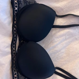 Svart push-up bh med spets och gulddetaljer från CNSRD - Svart push-up bh med vadderade kupor, spetsdetaljer på axelbanden och guldfärgade ringar samt mönster längs bandet. Snygga dubbla band framtill och dekorativa detaljer som ger en exklusiv känsla.