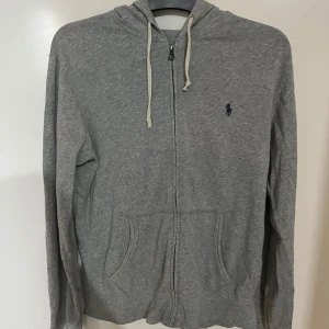 Grå hoodie från Polo Ralph Lauren - Grå hoodie från Polo Ralph Lauren med dragkedja framtill, huva med vita snören och klassisk blå logga på bröstet. Tillverkad i mjuk bomull och har två fickor framtill. Perfekt för en chill och stilren look.