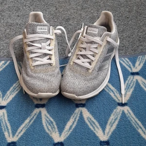 Gråvita Adidas sneakers med Cloudfoam - Snygga Adidas sneakers i grått och vitt med stickad look och silvriga ränder på sidorna. Skorna har vit sula med Cloudfoam-dämpning och klassisk snörning. Perfekta för dig som gillar sportig stil och vill ha bekväma skor till vardagen.