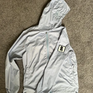 Season one OG!! Ljusblå vindjacka från Risky - Snygg ljusblå vindjacka från Risky med huva, dragkedja och broderad logga på huvan. Jackan har en patch på ärmen och är tillverkad i lätt polyester. Perfekt för dig som vill ha en clean och stilren look under sommaren