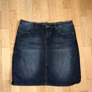 Mörkblå jeanskjol med broderade bakfickor - Säljer en klassisk mörkblå jeanskjol med snygga broderade detaljer på bakfickorna. Kjolen har normal passform, bälteshällor och knappstängning framtill. Är 172 och det går ungefär två tredje delar ner på mina lår.