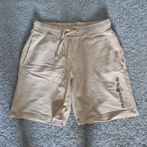 Beige shorts från Sail Racing med snörning - Beiga shorts från Sail Racing med snörning i midjan och broderad logga på ena benet. Mjukt material och en bakficka med dragkedja. Perfekta för varma dagar.