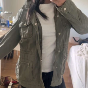 Olivgrön jacka med paljettvingar från MY T - Säljer en olivgrön jacka från MY T i storlek S. Jackan har långa ärmar, flera fickor framtill och snygga paljettvingar på ryggen som ger en unik look. Perfekt för dig som vill sticka ut lite extra.