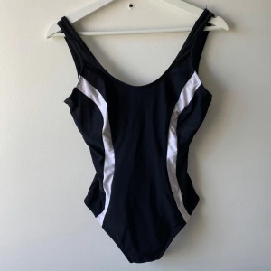 Svart baddräkt med vita detaljer - En sportig svart baddräkt med vita paneler längs sidorna. Djup rygg och inbyggd vit bh för extra stöd. 