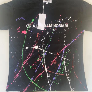 Maison margiela t shirt - Cool svart t-shirt från Maison Margiela MM6 med färgstänk i grönt, rosa, vitt, lila och blått samt tryckt text på framsidan. Rund halsringning och korta ärmar. Tillverkad i Italien.