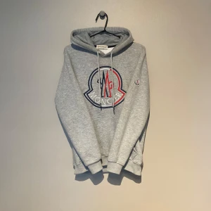 Grå hoodie från Moncler - Säljer en grå hoodie från Moncler med stor broderad logga i rött, blått och vitt på bröstet samt en mindre logga på ärmen. Stl M men sitter som S!
