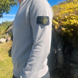 Grå sweatshirt från Stone Island - Säljer en stilren grå sweatshirt från Stone Island med klassisk patch på vänster ärm. Tröjan har rund halsringning och ribbade muddar vid ärmslut och nederkant. Perfekt för en avslappnad och snygg look.