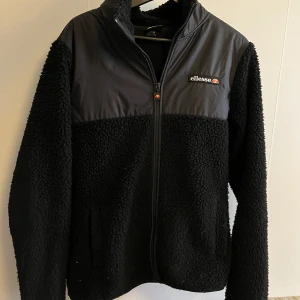 Svart fleecejacka från Ellesse - Säljer en svart fleecejacka från Ellesse med dragkedja framtill. Jackan har mjuk teddyfleece på ärmar och kropp samt slätare tyg upptill och på axlarna. Ellesse-logga på bröstet. Perfekt för kyliga dagar.