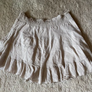 Beige volangkjol från Vila - Supersöt beige kjol från Vila med volangkant och smock i midjan. Kjolen är kort och har en luftig känsla, perfekt för varma dagar. Tillverkad i ett lätt material.