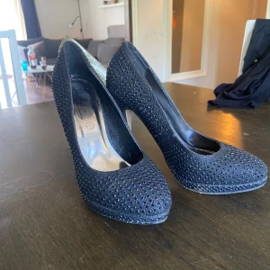 Svarta glittriga klackskor från Ruffle Collection - Säljer ett par svarta klackskor från Ruffle Collection med glittriga detaljer över hela skon. Skorna har hög klack, perfekta för att sticka ut lite extra. Klassisk och elegant design som passar till många tillfällen.