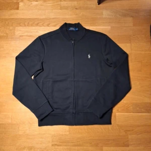 Svart tröja från Polo Ralph Lauren - Säljer en stilren svart tröja från Polo Ralph Lauren med dragkedja och broderad logga på bröstet. Tröjan har långa ärmar och är tillverkad i bomullsmaterial. Perfekt för en avslappnad och snygg look.