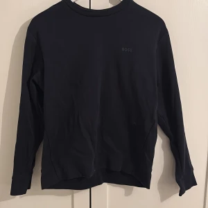 Blå tröja från BOSS - Säljer en stilren blå tröja från BOSS med diskret logga på bröstet. Tröjan har långa ärmar och en klassisk rund halsringning. Perfekt för en minimalistisk look. Tröjan är i bra skick och inköpt från Boss butiken i Malaga. Tröjan har en mörkblå färg.