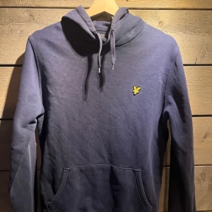 Mörkblå hoodie från Lyle & Scott - Säljer en snygg mörkblå hoodie från Lyle & Scott med deras ikoniska gula logga på bröstet. Tröjan har en klassisk känguruficka och justerbar huva med snören. Perfekt för en avslappnad stil.