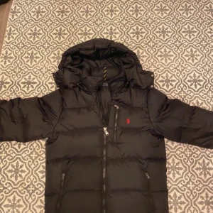 Svart dunjacka från Ralph Lauren - Säljer en snygg svart dunjacka från Ralph Lauren med huva och dragkedja. Jackan har en liten röd logga på bröstet och är perfekt för kyliga dagar. Den är långärmad och har en quiltad design för extra värme.