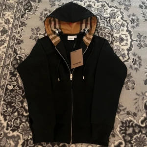 Svart huvtröja från Burberry - Säljer en stilren svart huvtröja från Burberry med dragkedja och den klassiska rutan i huvan. Perfekt för en casual look med en touch av lyx. Tröjan har långa ärmar och snörning vid huvan.