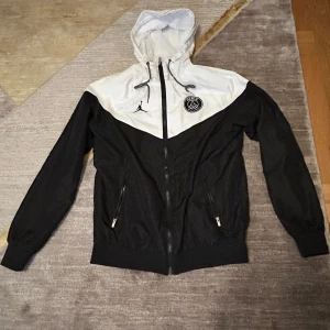 Svart och vit regn/vindjacka från Jordan x PSG - Säljer en snygg svart och vit vindjacka från Jordan x PSG. Jackan har en dragkedja framtill och en justerbar huva med snörning. Den är dekorerad med Jordan-loggan och PSG-emblemet på bröstet. Perfekt för sportiga tillfällen eller som en stilren vardagsjacka.. Använt max 1 gång , så den är helt ny 