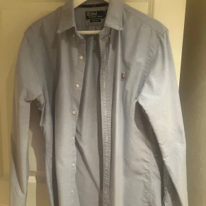 Ljusblå skjorta från Polo Ralph Lauren - Snygg ljusblå skjorta från Polo Ralph Lauren med klassisk krage och knappar framtill. Skjortan har en broderad logotyp på bröstet och är långärmad. Perfekt för en stilren look.