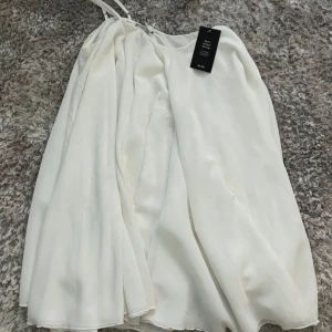 Studentklänning - Säljer en stilren vit studentklänning från NA-KD i storlek EU 36. Klänningen har tunna axelband och en luftig passform, perfekt för varma dagar. Den är tillverkad i ett lätt och flödande material som ger en elegant look.