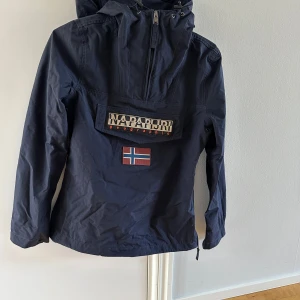 Mörkblå jacka från Napapijri i nyskick  - Säljer en mörkblå skaljacka från Napapijri med huva och dragkedja. Jackan har en stor ficka framtill med logotyp och norsk flagga. Perfekt för utomhusaktiviteter med stil. 🧥