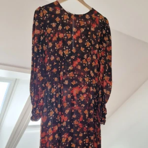 Blommig långklänning - Säljer en vacker långklänning med blommigt mönster i rött och orange på en mörk bakgrund. Klänningen har långa ärmar och en elegant passform som passar perfekt för höst och vår. Perfekt för dig som vill ha en stilren look med en touch av färg.