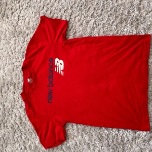 Röd t-shirt från New Balance - Säljer en röd t-shirt från New Balance med deras ikoniska logga i vitt och blått på framsidan. Perfekt för en sportig look eller avslappnad stil. Tillverkad i mjukt material för extra komfort.