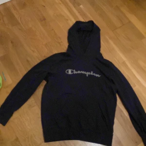 Svart hoodie från Champion - Säljer en mörkblå hoodie från Champion i storlek L. Den är tillverkad av 100% bomull och har en klassisk Champion-logga på bröstet. Perfekt för en avslappnad stil.