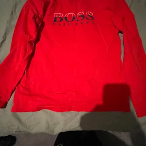 Röd långärmad tröja från Hugo Boss - Säljer en snygg röd långärmad tröja från Hugo Boss med logotypen tryckt på framsidan. Perfekt för en stilren och avslappnad look. Passar bra till både jeans och kjol.