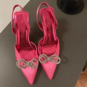 Rosa pumps med klack och rosett - Snygga rosa pumps i satin med spetsig tå och elegant klack. Skorna har en dekorativ rosett framtill som ger en extra touch av glamour. Perfekta för en festlig kväll!