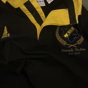 AIK retro tröja - Säljer en svart fotbollströja från Adidas med gula detaljer och AIK-logga. Tröjan har en gul krage och texten 'Råsunda Stadion 1937-2012' tryckt på framsidan. Väldigt unik men för liten storlek M