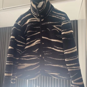 Svart och beige teddyjacka med zebramönster - Säljer en cool teddyjacka i svart och beige med zebramönster. Jackan har en dragkedja framtill och hög krage för extra värme. Perfekt för att hålla stilen i kyligare väder! Slutsåld 