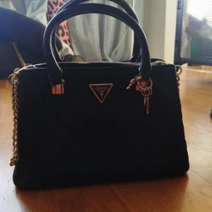 Svart handväska från Guess - Snygg svart handväska från Guess med gulddetaljer och kedjeaxelrem. Väskan har en dragkedja på baksidan och ett triangulärt Guess-emblem på framsidan. Perfekt för att ge en elegant touch till din outfit. Aldrig använd! Nypris 2000:-