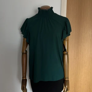Mörkgrön blus med volangdetaljer - Snygg mörkgrön blus med hög krage och volangdetaljer på ärmarna. Använt ett fåtal gånger.