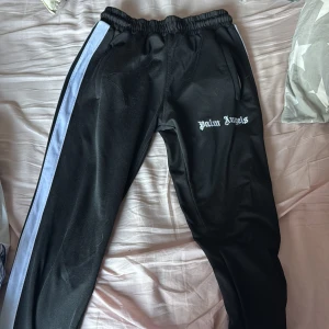 Svarta träningsbyxor från Palm Angels - Snygga svarta träningsbyxor från Palm Angels med vita sidoremsor och elastisk midja. Byxorna har en broderad logga på sidan och är tillverkade i polyester för en bekväm passform. Qr kod till hemsidan.! Priset kan diskuteras 