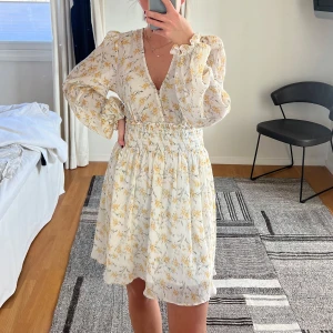 Blommig klänning med v-ringning - Charmig blommig klänning från NA-KD med v-ringning och långa ärmar. Vit med gula blommor och lite grönt. Den har en elastisk midja. Perfekt för vår och sommar med sitt lätta och luftiga material.