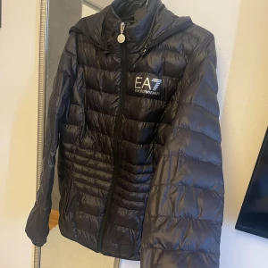Armani jacka svart - Säljer en snygg, svart dunjacka från Emporio Armani med EA7-logga på bröstet. Luvan på jackan går att ta bort och sätta på. Jackan har en liten deffekt frampå ( bild 4 ). Ena dragkedjan på fickan har lossnat men det går fortfarande att stänga fickan som vanligt. Utöver det är jackan i bra skick.