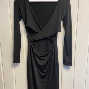 Svart omlottklänning - Snygg svart omlottklänning med lång ärm och v-ringad design. Klänningen har en unik öppen detalj och är perfekt för en elegant look.