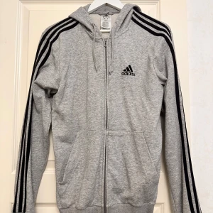Grå hoodie från Adidas - Säljer en grå hoodie från Adidas med dragkedja och svarta ränder längs ärmarna. Den har en justerbar huva med snören och det klassiska Adidas-loggan på bröstet. Perfekt för en sportig look! Snören saknar plast