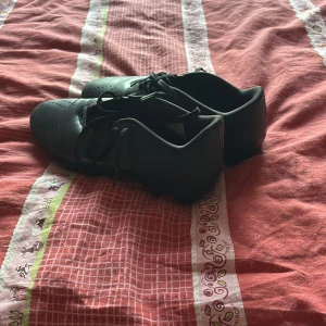 Svarta fotbollsskor från Adidas - Säljer ett par svarta fotbollsskor från Adidas med snörning och dobbar för optimalt grepp på planen. Perfekta för fotbollsträning och matcher. Skorna har en stilren design och är tillverkade i syntetmaterial.