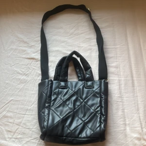 Svart quiltad axelväska från H&M Divided - Säljer en snygg svart quiltad axelväska från H&M Divided. Väskan har både handtag och en justerbar axelrem för flexibel användning. Perfekt för att bära dina dagliga nödvändigheter med stil.