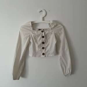 Vit blus med knappar - Säljer en söt vit blus med långa ärmar och knappar framtill. Blusen har en croppad passform och elastiska ärmslut. Perfekt för en stilren look.