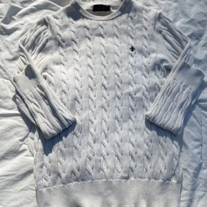 Vit cabbelstickad tröja från Morris - Säljer en stilren vit cabbelstickad tröja från Morris med ett diskret broderat emblem på bröstet. Tröjan har ett klassiskt flätmönster och långa ärmar. Perfekt för en elegant och avslappnad look. Tröjan är i bra skick. Skriv till mig vid frågor eller om du vill ha fler bilder.