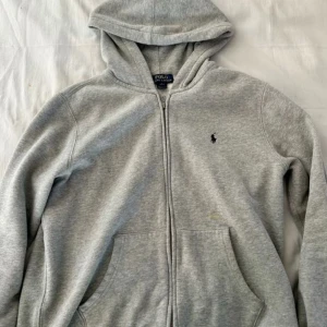 Grå hoodie från Polo Ralph Lauren - Säljer en klassisk grå hoodie från Polo Ralph Lauren med dragkedja och huva. Tröjan har långa ärmar och en liten broderad logga på bröstet. Perfekt för en avslappnad stil.