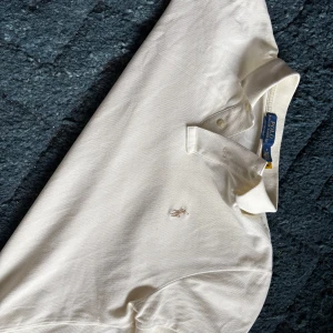 Vit pikétröja från Ralph Lauren - Säljer en klassisk vit pikétröja från Ralph Lauren med korta ärmar och den ikoniska loggan broderad på bröstet. Perfekt för en stilren och avslappnad look. Tröjan har en krage med knappar för en tidlös stil.