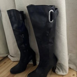 Svarta stövlar med spänne - Snygga svarta stövlar i skinn med hög klack och dekorativt spänne upptill. De har en dragkedja på sidan för enkel på- och avtagning. Perfekta för att ge en stilren touch till din outfit.