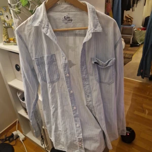 Ljusblå randig skjorta från Lee (over size) - Säljer en stilren ljusblå skjorta från Lee med diskreta ränder och knappar framtill. Skjortan har två bröstfickor och långa ärmar, perfekt för en avslappnad look. Passar bra till både jeans och chinos. 🕶️ eller en kort snygg kjol / jeans shorts på sommaren . Nyskick. Storlek (slim) /oversize style